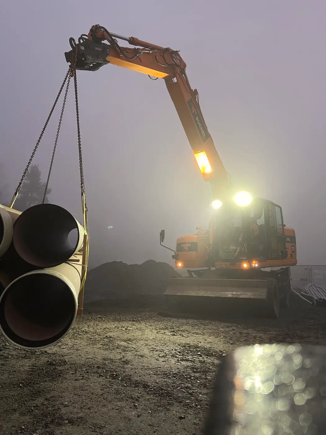 Bagger hebt im Morgennebel bei Dunkelheit zwei große Stahlrohre an