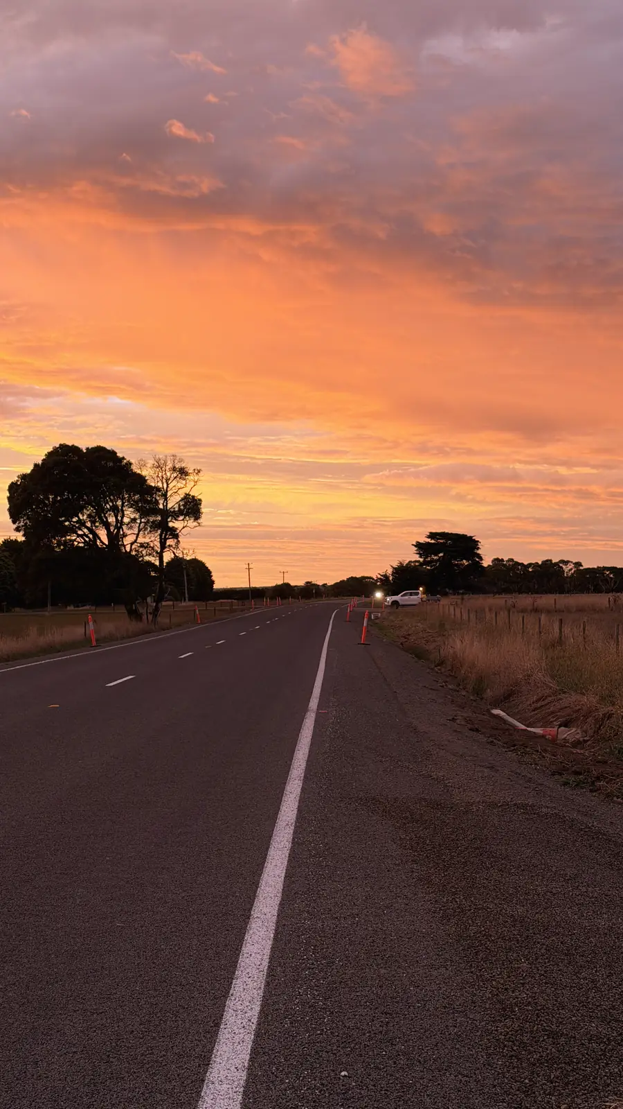 Landstraße bei Sonnenaufgang unter einem orange gefärbten Himmel, am Rand ein Pick-up mit eingeschalteten Scheinwerfern