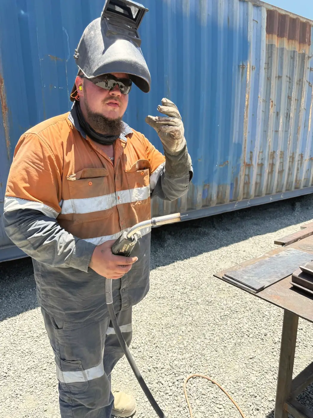 Marvin in oranger Warnkleidung mit hochgeklapptem Schweißerhelm vor einem Container