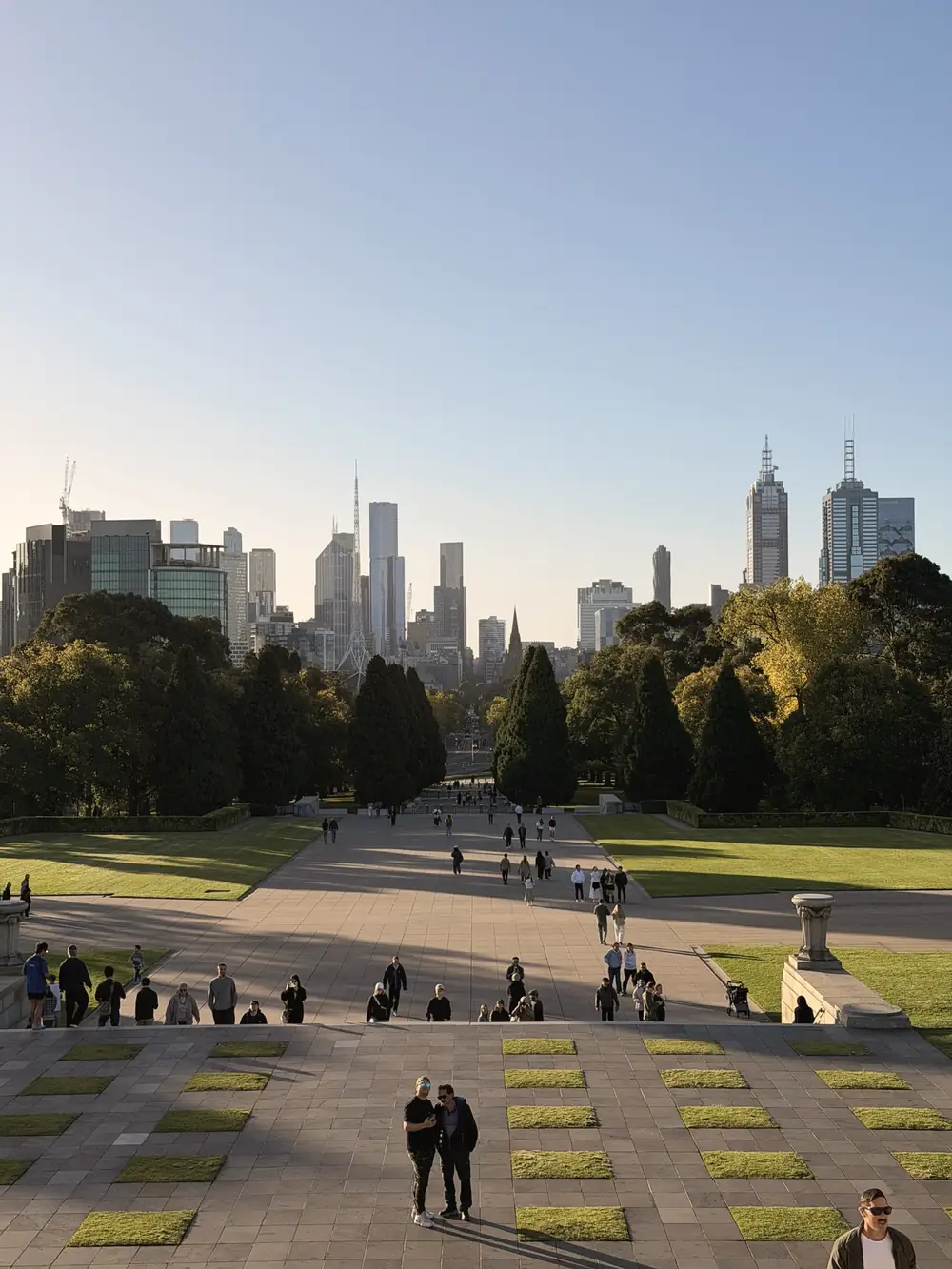 Skyline von Melbourne über einem weiten Park, davor Leute auf den Stufen