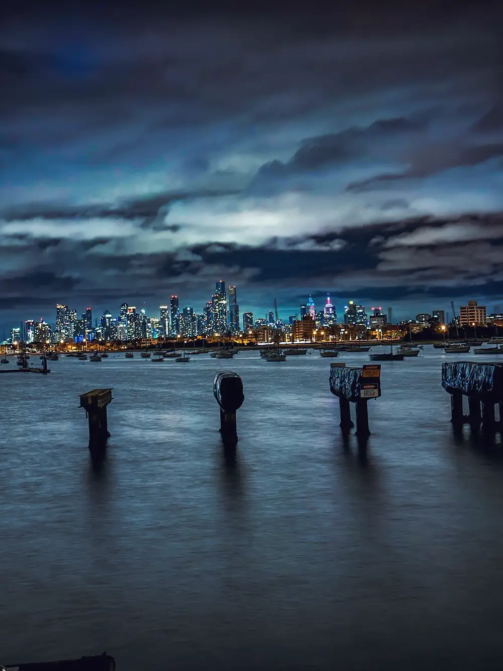 Skyline von Melbourne bei Nacht, davor alte Pfähle im Wasser