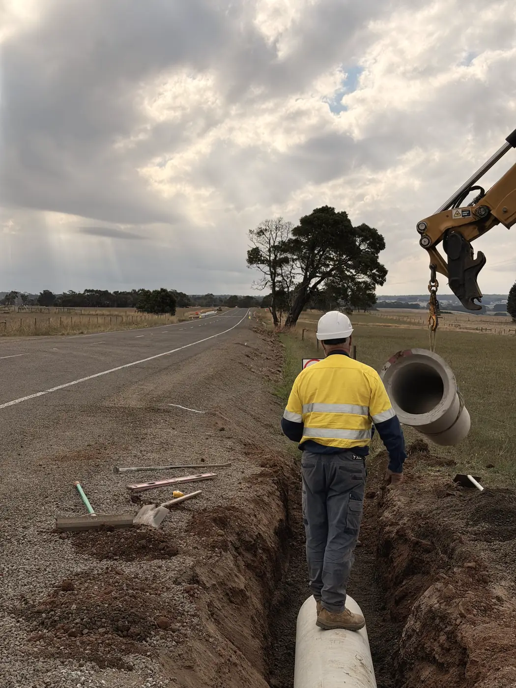 Arbeiter läuft in einem offenen Graben neben verlegten Betonrohren, Sonnenstrahlen brechen durch Wolken